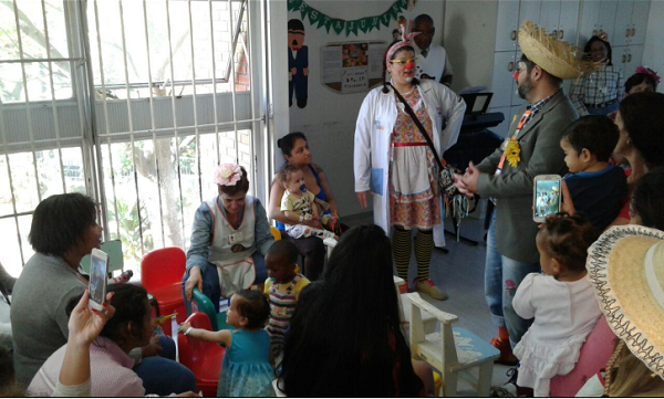 #PraCegoVer: Voluntários trajados com vestimentas de palhações tocam instrumentos para as crianças ao redor, que brincam com brinquedos próximos